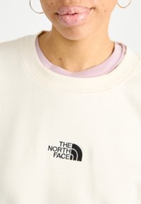 Sweatshirt crème avec le logo "THE NORTH FACE" brodé en noir. Superposé avec une chemise violet clair visible au niveau du col.
