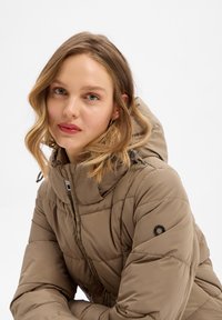 Braune Puffjacke mit Kapuze, gestepptem Design, Vorderreißverschluss und verstellbarem Bund. Mit schwarzen Akzenten an den Ärmeln und silbernten Beschlägen.