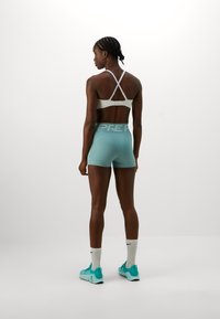 Lyseblå treningsshorts med bred linning som har teksten "PRE". Modellen har på seg en hvit sports-bh med kryssrygg og tealfargede joggesko.