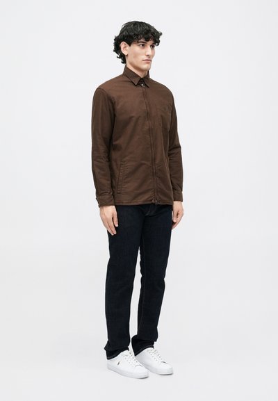 Camisa castanha com fecho éclair feita de um tecido suave, com colarinho ponto e bolsos laterais. Combinada com calças de ganga escuras e sapatilhas brancas.