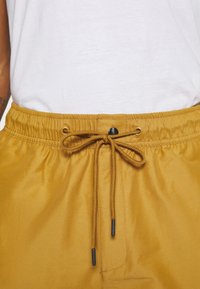 Žluté kraťasy s elastickým pasem a šňůrkou. Mají hladkou texturu a černý knoflík na přední straně.