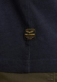 Navy cotton t-shirt featuring a PME Legend label with yellow text and wing graphic, stitched at the lower corner against a textured background.