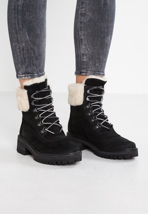 Bottes d'hiver en suède noir avec doublure en fourrure blanche, œillets en métal et semelles épaisses à crampons, portées avec un jean noir délavé.