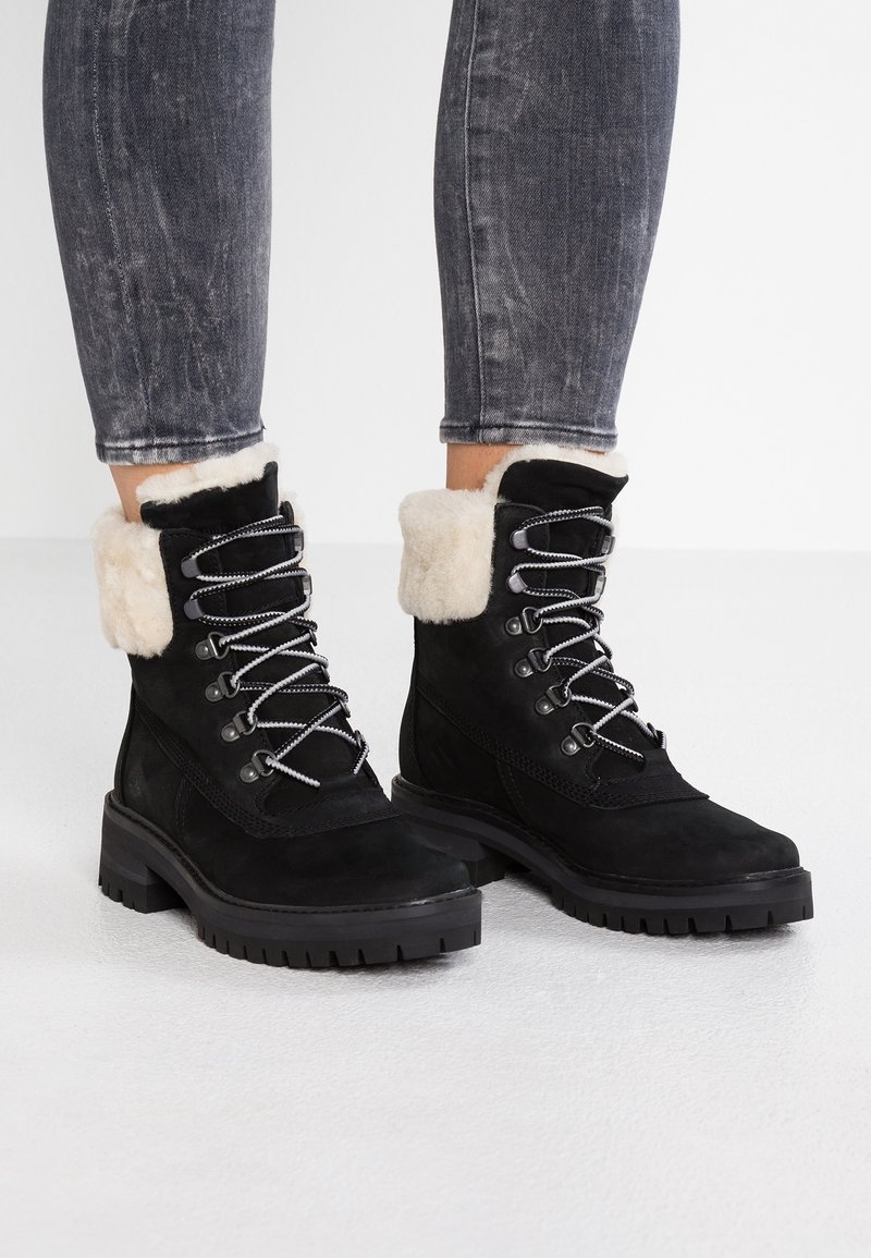 Bottes d'hiver en suède noir avec doublure en fourrure blanche, œillets en métal et semelles épaisses à crampons, portées avec un jean noir délavé.