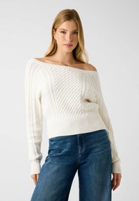 Pull en maille torsadée blanc avec design à épaules dénudées et ourlet côtelé, associé à un pantalon en denim bleu, présentant une coupe décontractée et un motif texturé.