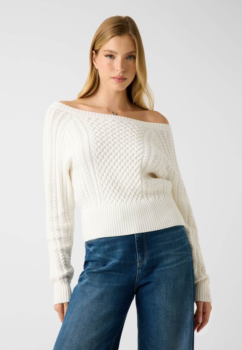 Pull en maille torsadée blanc avec design à épaules dénudées et ourlet côtelé, associé à un pantalon en denim bleu, présentant une coupe décontractée et un motif texturé.
