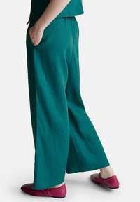 Persona che indossa pantaloni larghi turchesi con vita elastica e scarpe magenta, in piedi con le mani nelle tasche su uno sfondo bianco.