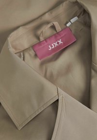 Camicia in tessuto beige, morbido al tatto, con colletto, dotata di un'etichetta rosa con la scritta "JJXX". Cuciture centrali e dettagli visibili.