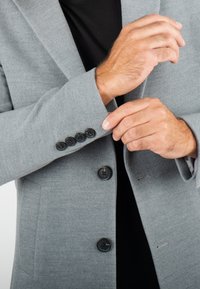 Homme portant un manteau gris en train de boutonner le poignet avec quatre boutons noirs, vêtu de noir en dessous, gros plan sur les mains et les détails du manteau.