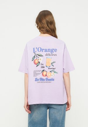 Person with long hair wearing a lavender t-shirt with colorful orange fruit and juice graphics and text on the back, paired with blue jeans.