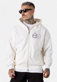 Haut de sweat à capuche blanc zippé en coton doux, avec un logo brodé gris sur la poitrine et des poignets côtelés. Porté par-dessus un t-shirt blanc.