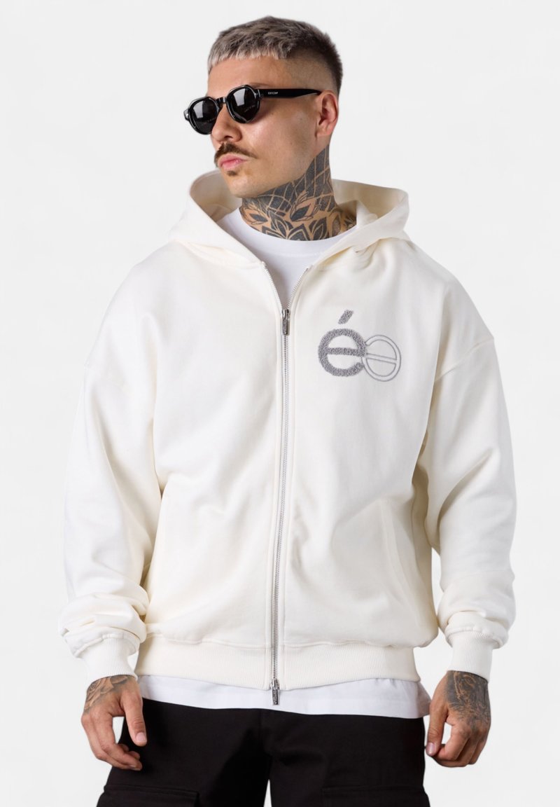 Weiße Zip-Hoodie aus weichem Baumwollstoff mit einem grauen gestickten Logo auf der Brust und gerippten Bündchen. Wird über einem weißen T-Shirt getragen.