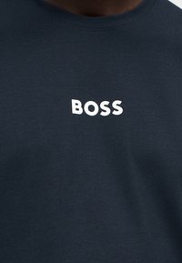 Σκούρο ναυτικό πουκάμισο με λευκά έντονα κεφαλαία γράμματα "BOSS" στο κέντρο του στήθους, φορεμένο από άτομο με σκουρόχρωμο δέρμα.