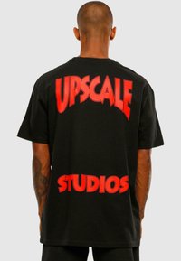 Čierne bavlnené tričko s veľkým červeným textom "UPSCALE" nad "STUDIOS" na zadnej strane. Krátke rukávy a voľný strih.