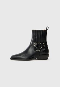 Botas de cuero negro hasta el tobillo con punta afilada, detalles acanalados en la parte trasera y una correa ancha con un hebilla circular y adornos de tachuelas.