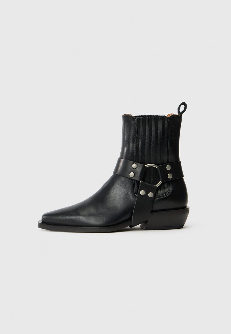 Botas de cuero negro hasta el tobillo con punta afilada, detalles acanalados en la parte trasera y una correa ancha con un hebilla circular y adornos de tachuelas.