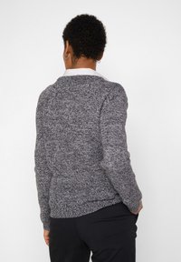 Personne aux cheveux courts et bouclés, portant des boucles d'oreilles anneaux et un pull gris chiné par-dessus une chemise blanche à col, vue de dos avec les mains dans les poches.