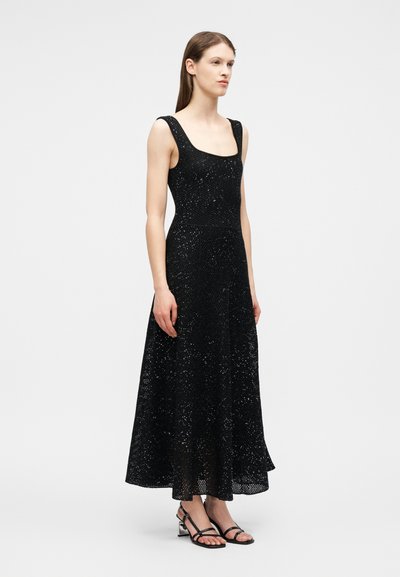 Γυναίκα που φοράει μαύρο μίντι φόρεμα με παγιέτες χωρίς μανίκια, συνδυασμένο με μαύρα σανδάλια με λουράκια και χοντρό τακούνι, στέκεται μπροστά σε απλό φόντο.