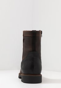 Ruskea nahkainen nilkkasaapas, jossa musta kumipohja, teksturoitu pinta, takasilmukka ja sivuvetoketju, esittelee kontrastista muotoilua.