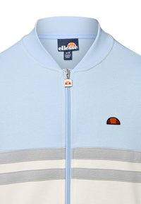 Lichtblauwe zip-up jack met een staande kraag, voorzien van grijze en witte horizontale strepen, en een herkenbaar geborduurd logo op de borst.