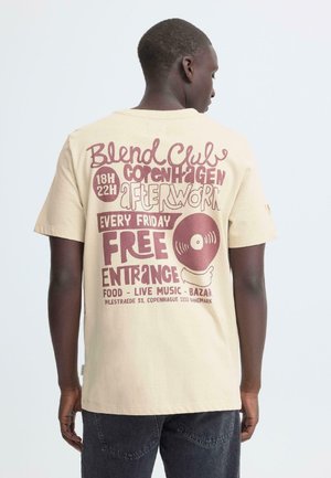 Beige katoenen t-shirt met rode tekstgrafieken op de achterkant die een evenement promoten, met een illustratie van een vinylplaat en diverse details.