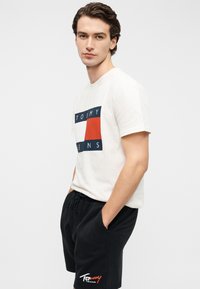 Λευκό βαμβακερό t-shirt με έντονο ναυτικό, κόκκινο και λευκό γραφικό λογότυπο· συνδυασμένο με μαύρο σορτσάκι που φέρει κόκκινο και λευκό κεντημένο λογότυπο στο πόδι.