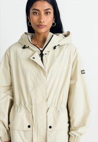 Világos bézs színű, kapucnis dzseki, elasztikus derékkal, két elülső zsebbel és húzózsinóros kapucnival. Simaképernyős, könnyű anyagból készült.