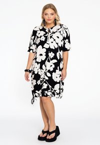 Zwarte jurk met grote witte bloemenprint, korte mouwen, geknoopte zoomdetail en een keyhole halslijn. Gecombineerd met zwarte platform sandalen.
