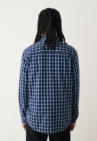 Chemise à carreaux bleue à manches longues, col boutonné et ourlet arrondi. Le tissu présente un motif à carreaux dans différentes nuances de bleu.