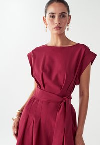 Žena s spiatymi vlasmi má na sebe bezrukávové červené šaty so zaväzovacím opaskom a zlaté a perlové visiacé náušnice na jednolatom pozadí.