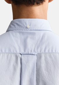 Vue de dos d'une personne portant une chemise bleu clair à col avec un détail bouton et pli sur le haut du dos.