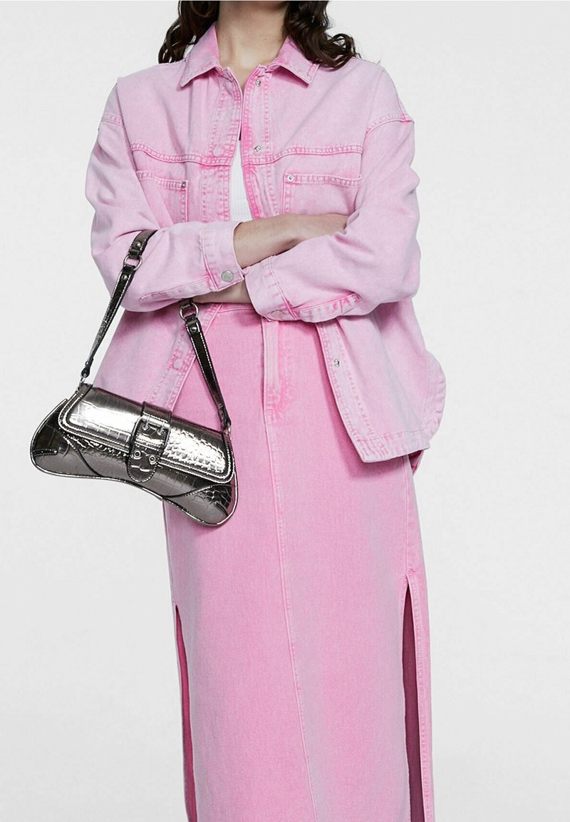 Femme portant une veste en jean rose et une jupe assortie avec des fentes sur les côtés, tenant un petit sac à main argenté brillant sur son bras.