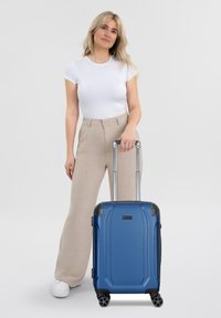 Maleta azul con un diseño rígido, vertical, mango telescópico y cuatro ruedas giratorias. La parte frontal presenta líneas en relieve y detalles en las esquinas.