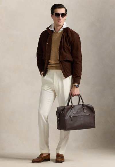 Homme portant une veste marron foncé, un pull beige, un pantalon blanc, des chaussures marron et des lunettes de soleil tenant un grand sac en cuir marron foncé.