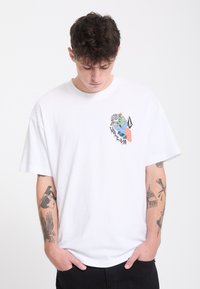 Jeune homme avec des tatouages sur les deux avant-bras regarde vers le bas, portant un t-shirt blanc avec un graphique coloré sur le côté gauche de la poitrine.