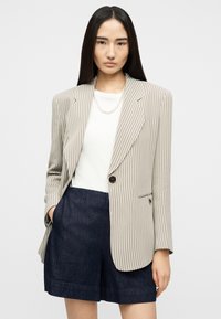 Mujer con cabello largo y oscuro, vestida con un blazer beige con rayas, blusa blanca, collar de perlas y pantalones cortos azul oscuro, posando frente a un fondo liso.