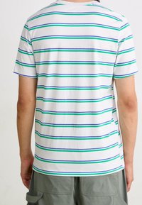 Bílé tričko s krátkým rukávem a kulatým výstřihem, které má vodorovné pruhy v zelené a modré barvě. Měkký bavlněný materiál s volným střihem.