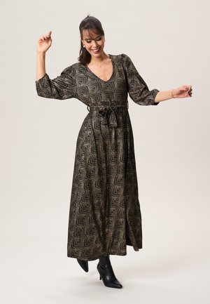 Vrouw lacht met opgeheven armen, draagt een lange jurk met patroon en een tailleband, en zwarte enkellaarzen. Ze staat tegen een effen achtergrond.