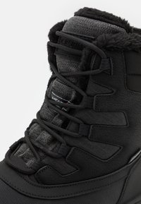 Bota aislante negra con tejido texturizado, forro de piel sintética, suela de goma y diseño de cordones con ojales reforzados y detalle de logo.