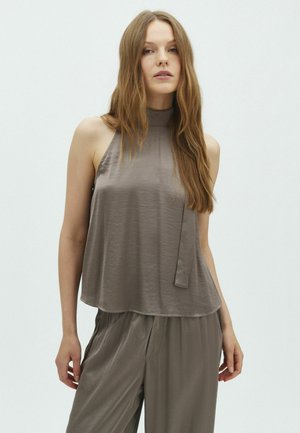 Femme aux longs cheveux bruns portant un haut taupe sans manches à col montant et un pantalon ample assorti, debout devant un fond clair uni.