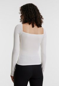 Langärmliges Oberteil aus weißem geripptem Material, mit quadratischem Rückenausschnitt und figurbetonendem Design, kombiniert mit schwarzen Hosen.
