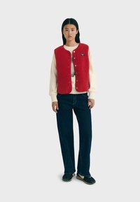 Rode fleece vest met zilveren drukknoopjes, gedragen over een crèmekleurig sweatshirt, gecombineerd met donkere denim broek en zwarte schoenen. Effen achtergrond.