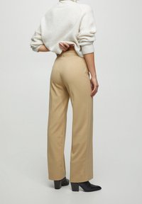 Pantalon beige à jambes larges en tissu lisse, avec une taille haute et des poches arrière, associé à un pull crème texturé et des bottes à talons noires.