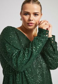 Vihreä paljettimekko, jossa on V-pääntie, pitkät hihat ja napilliset hihansuut. Oma teksturoitu pinta ja heijastavat paljetit luovat kimaltavan vaikutelman.