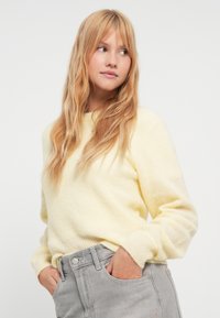 Κίτρινο πουλόβερ από μαλακό, υφασμένο ύφασμα με στρογγυλή λαιμόκοψη και ριμπ μανσέτες, σε συνδυασμό με γκρι παντελόνι από τζιν με κλείσιμο με κουμπιά.