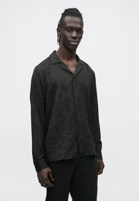 Moški s tkanimi lasmi, oblečen v črno srajco s potiskom in črne hlače, stoji pred enobarvnim svetlim ozadjem.