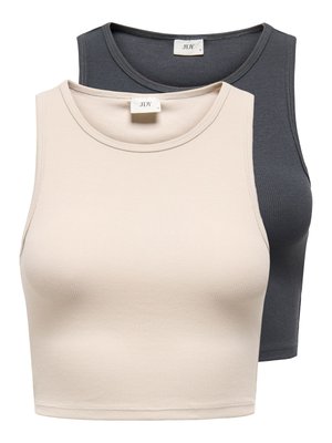 Vrouw die een lichtbeige pet draagt, een donkergrijze mouwloze cropped top, zwarte skinny jeans met riem en witte sneakers, staand tegen een effen lichte achtergrond.