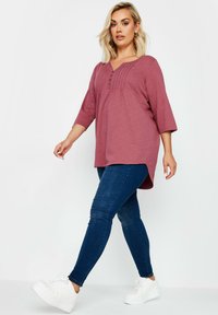 Burgundin tunika, jossa on kolmen neljänneksen hihat ja poimutus yksityiskohtina, yhdistettynä tummansinsisiin skinny-farkkuihin ja valkoisiin lenkkareihin.