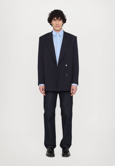 Blazer bleu marine à double boutonnage avec deux grands boutons, chemise bleu clair, jeans droits noirs et chaussures noires. Design simple et structuré.