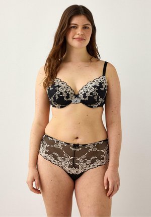 Nainen, joka pukeutunut mustaan pitsirintaliiveihin ja niihin sopiviin korkeavyötäröisiin pitsialushousuihin, seisoo vaaleaa taustaa vasten katsellen eteenpäin.